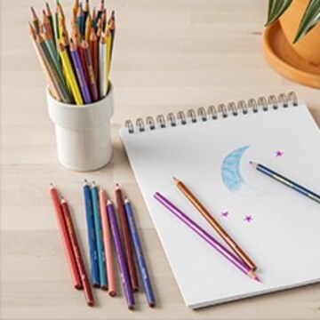 colored pencils in a cup, on a wood like table, and on a paper pad. There is also the begining of a moon drawing with the moon filled in, in blue and some stars in purple